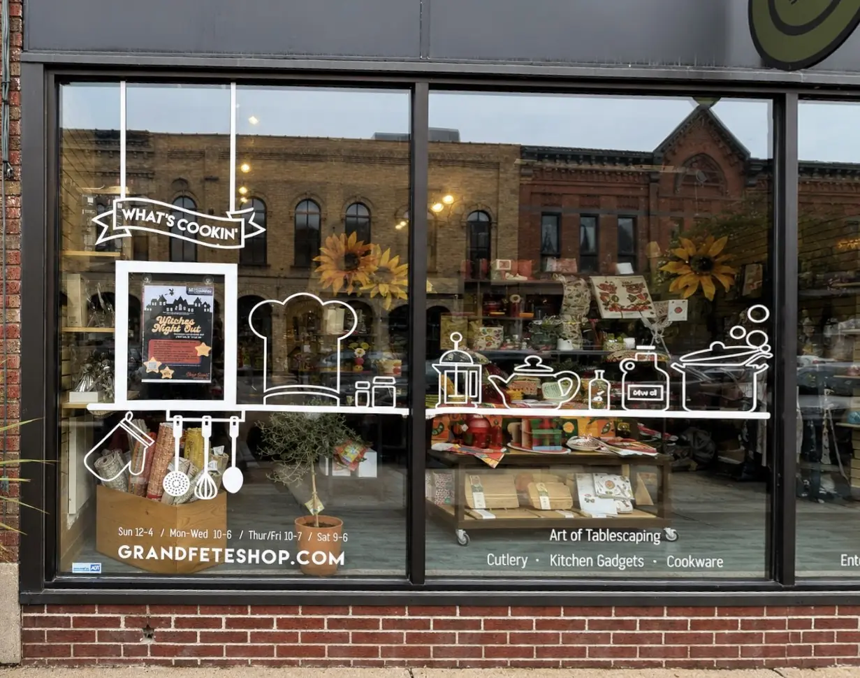 A kitchen shop window drawn with white line illustrations of utensils and cookware under a What's Cookin' banner, with the shop's hours and website.